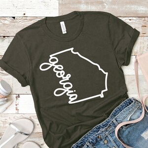 May include: A dark green t-shirt with a white outline of the state of Georgia and the word "Georgia" written in a cursive font.