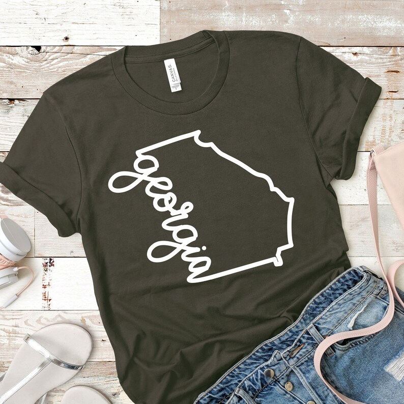 May include: A dark green t-shirt with a white outline of the state of Georgia and the word "Georgia" written in a cursive font.