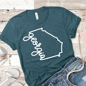 May include: Teal blue t-shirt with a white outline of the state of Georgia and the word "Georgia" written in a cursive font.