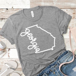 May include: A gray t-shirt with a white outline of the state of Georgia and the word "Georgia" written in a cursive font.