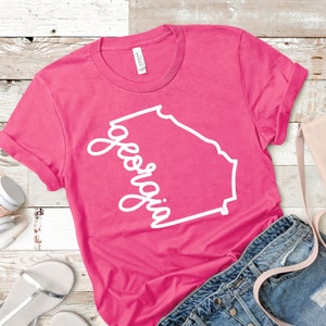 May include: Pink t-shirt with a white outline of the state of Georgia and the word "Georgia" written in a cursive font.
