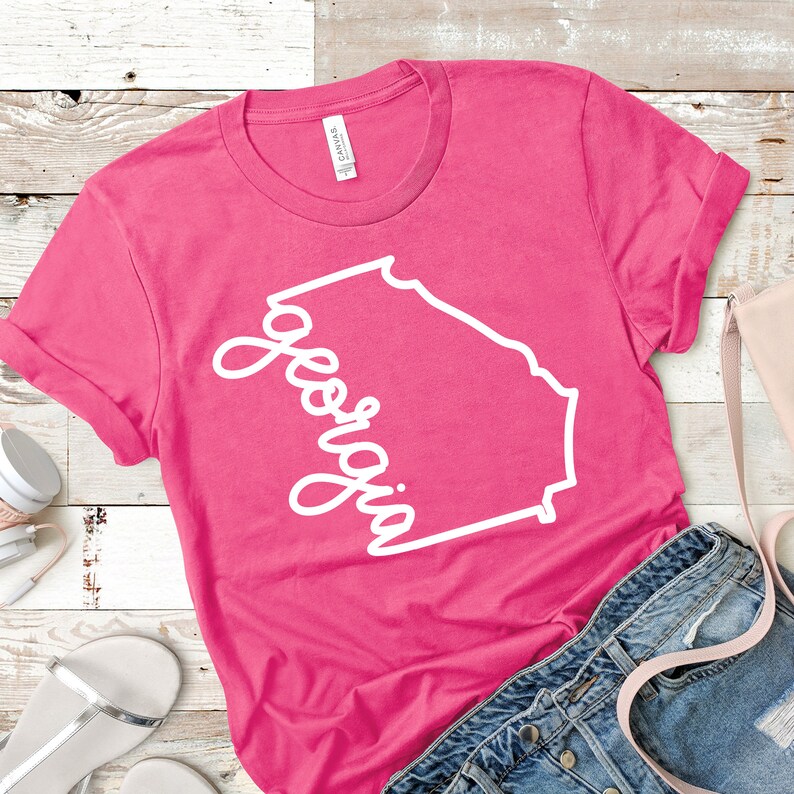 May include: Pink t-shirt with a white outline of the state of Georgia and the word "Georgia" written in a cursive font.