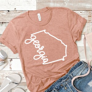 May include: A peach-colored t-shirt with a white outline of the state of Georgia and the word "Georgia" written in a cursive font.