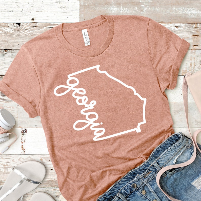 May include: A peach-colored t-shirt with a white outline of the state of Georgia and the word "Georgia" written in a cursive font.