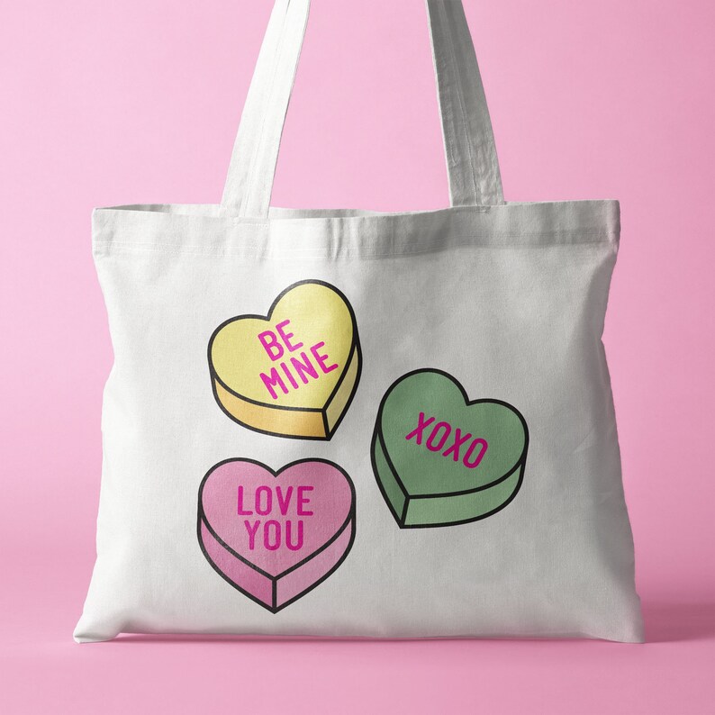 Pu&ograve; includere: Borsa tote bianca con tre caramelle a forma di cuore con testo. La caramella gialla dice "BE MINE", la caramella verde dice "XOXO" e la caramella rosa dice "LOVE YOU". La borsa ha due manici bianchi.