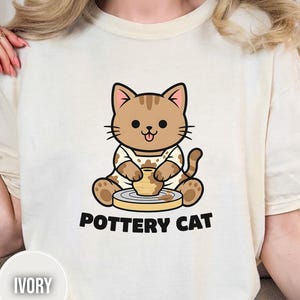 May include: Ivory-colored t-shirt featuring a cartoon cat creating pottery. The cat is brown with a white shirt and is sitting at a pottery wheel. The words "POTTERY CAT" are printed below the cat.