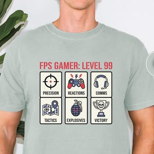 May include: A pale green t-shirt featuring the text "FPS GAMER: LEVEL 99" and six gaming-related icons. The icons include a target, controller, headphones, map, grenade, and trophy. The shirt is a casual style.