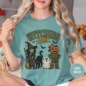 May include: A light green t-shirt featuring a graphic print with the text "WITCHING HOUR" in a stylized font. The design includes a green-skinned witch holding a broom, a black cat, a small white ghost, and a pumpkin-headed scarecrow figure.