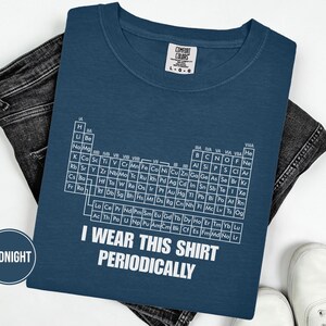 Puede incluir: Una camiseta azul marino con el texto "I WEAR THIS SHIRT PERIODICALLY" y un diseño de tabla periódica blanca. La camiseta está doblada sobre un par de vaqueros oscuros y zapatillas blancas.