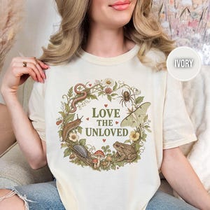 May include: An ivory t-shirt featuring a circular botanical wreath graphic with the text LOVE THE UNLOVED. The design includes illustrations of a snake, spider, moth, toad, slug, and mushrooms in muted earth tones with small red heart accents.
