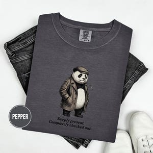 May include: A dark grey Comfort Colors t-shirt with a cartoon panda wearing a cap and coat. The panda has a thoughtful expression. The text "Deeply present. Completely checked out." is below the panda. The shirt is styled with jeans and white trainers.