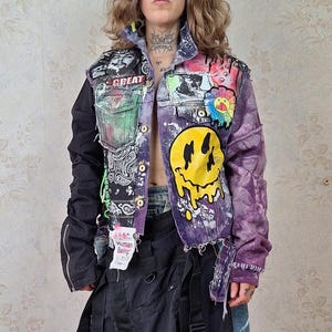 Op de afbeelding: Een veelkleurig spijkerjack met een grote gele smeltende smiley-graphic, diverse patches en tekst, gecombineerd met een lange zwarte rok met gespriemen en blauwe denim zijpanelen, en bruine schoenen van getextureerd leer.