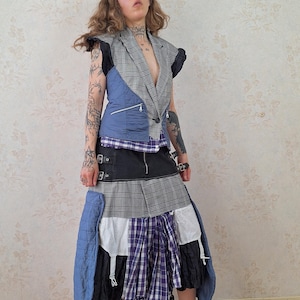Peut inclure: Un gilet sans manches à motif écossais gris et panneaux matelassés bleus, associé à une jupe superposée composée de tissu noir, sections écossaises grises, un panneau à carreaux violets et blancs et tissu blanc, avec des chaussures en cuir marron.