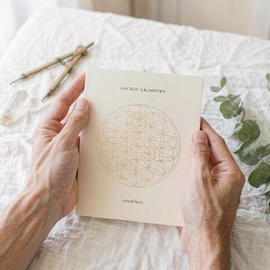 Pode incluir: Um diário de cor creme com um padrão geométrico estampado em folha de ouro no centro. O texto SACRED GEOMETRY está impresso acima do design e a palavra JOURNAL está impressa abaixo em uma fonte sem serifa simples e escura.