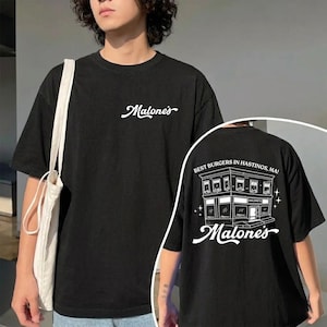 May include: Black t-shirt with the word "Malone's" in white script on the front. The back of the shirt features a white illustration of a building with the text "Best Burgers in Hastings, MA!" and "Malone's".