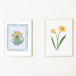 Puede incluir: Dos impresiones artísticas enmarcadas. Una presenta una tetera verde llena de flores coloridas, sobre un fondo azul claro con un borde festoneado. La otra impresión muestra un narciso amarillo con hojas verdes.