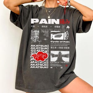 May include: Dark gray t-shirt with the word "PAIN" in white, along with Japanese characters and graphic elements. The shirt features the text "AKATSUKI" and a red cloud design. The shirt has a vintage, worn look.