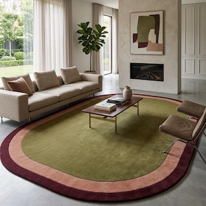 May include: An oval area rug featuring a large olive green center surrounded by a wide dusty pink border and an outer dark burgundy trim. The rug is placed on a smooth concrete floor in a living room setting with a sofa and chairs nearby.