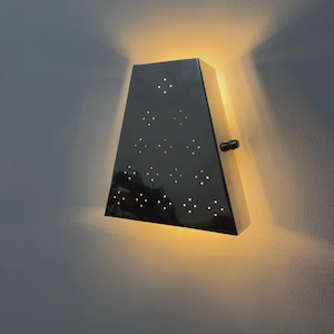 May include: A black, geometric wall sconce with star-shaped cutouts. The sconce is lit from within, emitting a warm glow. It has a small, round handle on the side. The design is modern.