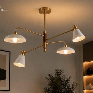 May include: A brushed brass chandelier featuring four adjustable arms with white conical and dome-shaped shades. The central column includes a dark wood accent. The text FINISH: Brushed Brass is visible in the upper left corner of the image.