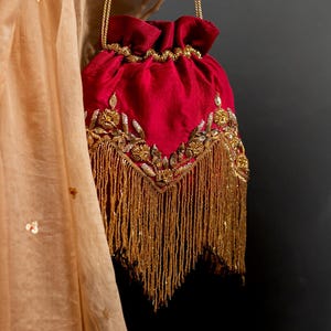 Puede incluir: Un bolso rojo con cordón, bordados y flecos dorados, colgado de una cadena dorada. El bolso está adornado con intrincados detalles dorados y una borla. Una tela color melocotón está drapeada en el fondo.