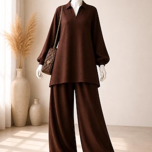 May include: A dark brown long-sleeved tunic with a collared V-neckline and matching wide-leg trousers. The outfit is paired with a small, dark brown quilted rectangular shoulder bag featuring a gold chain strap and light-colored pointed shoes.