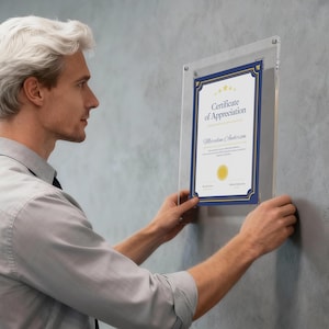 May include: A clear acrylic certificate frame with a blue and gold certificate inside. The certificate reads "Certificate of Appreciation" with a gold seal and decorative border. The frame is being held against a gray wall.