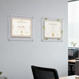 May include: Two clear acrylic certificate frames mounted on a gray wall. Each frame holds a certificate with gold and beige accents. One certificate is plain, the other has floral embellishments.