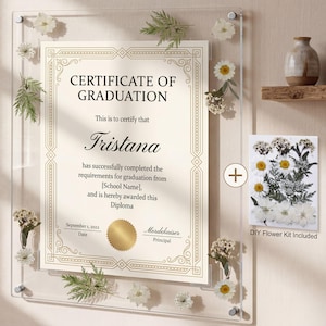 May include: A clear acrylic frame holds a graduation certificate with the name Tristana. The certificate is decorated with gold accents and floral embellishments. A DIY flower kit is included, along with a small vase on a wooden shelf.