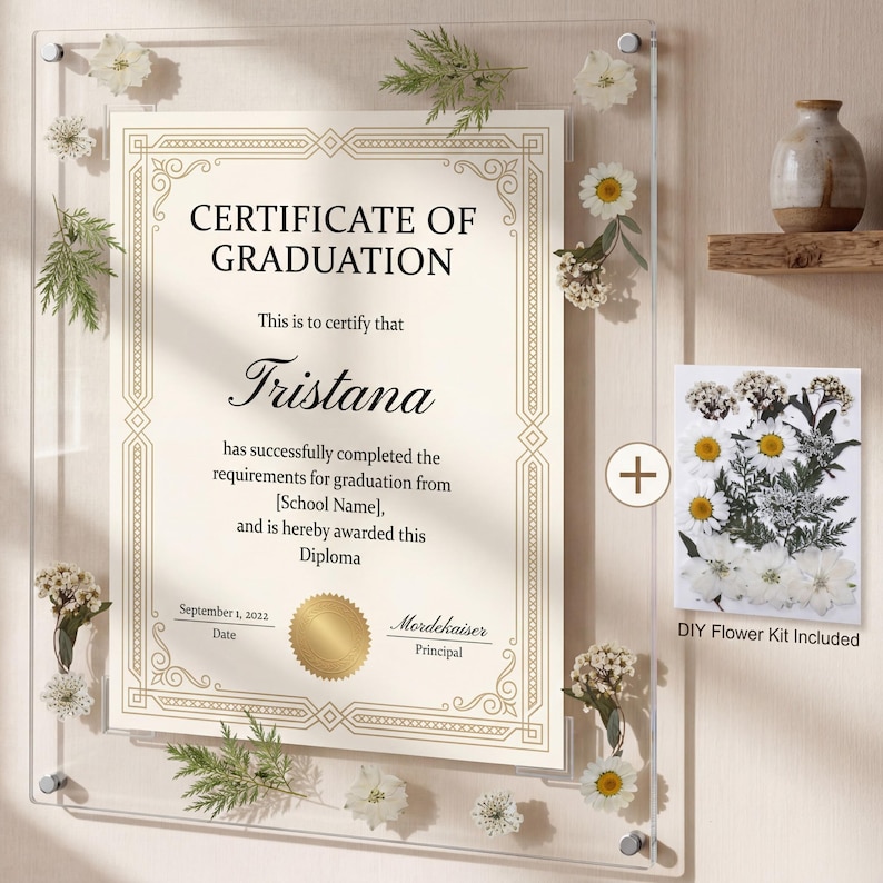May include: A clear acrylic frame holds a graduation certificate with the name Tristana. The certificate is decorated with gold accents and floral embellishments. A DIY flower kit is included, along with a small vase on a wooden shelf.
