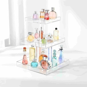 Peut inclure: Un pr&eacute;sentoir &agrave; trois niveaux en acrylique transparent avec une base carr&eacute;e et deux &eacute;tag&egrave;res carr&eacute;es plus petites empil&eacute;es verticalement. Le support contient divers flacons de parfum en verre de formes, tailles et couleurs vari&eacute;es, dont rose, bleu et dor&eacute;.