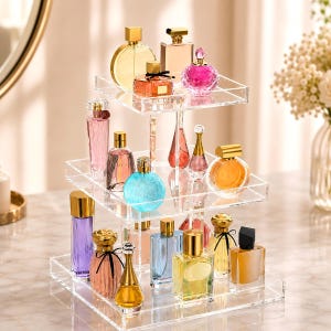 May include: A three-tiered clear acrylic display stand holding various glass perfume bottles in assorted shapes, sizes, and colors including pink, blue, yellow, purple, and orange, featuring gold and silver caps on a marble-patterned surface.