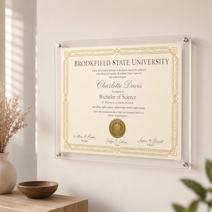 May include: A framed diploma from Brookfield State University, displayed on a white wall. The diploma is for a Bachelor of Science in Business Administration, with gold accents and text. The frame is clear acrylic, and the diploma is secured with silver hardware.