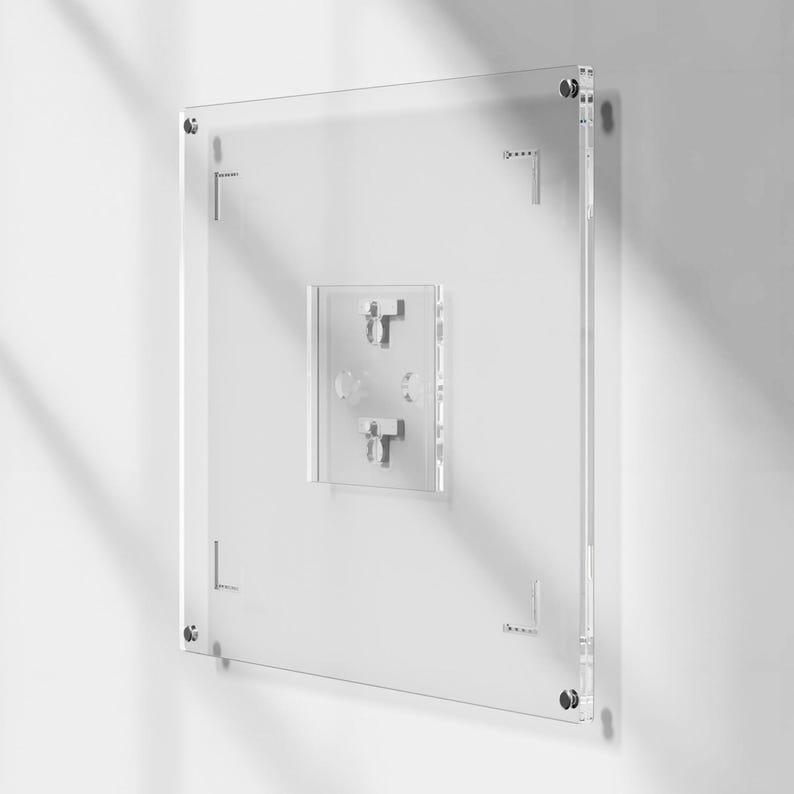 May include: A clear acrylic wall-mounted display case with a square shape. The case has a smaller, rectangular section in the center with two metal hooks. Metal hardware is visible at each corner and along the sides for mounting. The display case is mounted on a white wall.