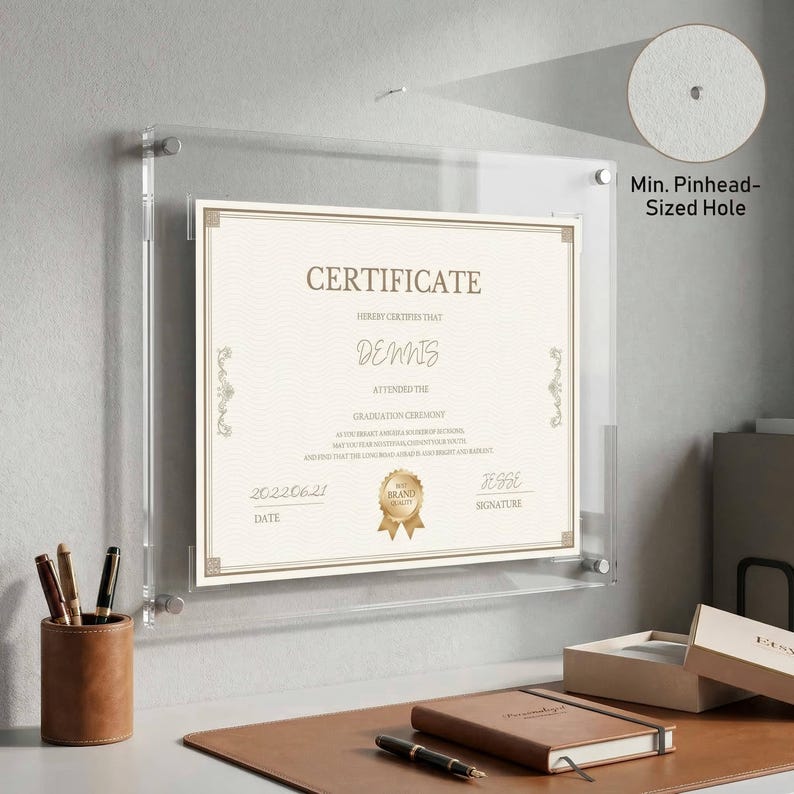 May include: A clear acrylic certificate frame displaying a certificate with the name "DENNIS" and the date "20220621". The frame is mounted on a gray wall. A leather pen holder and a notebook are visible on a desk.