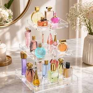 Peut inclure: Un pr&eacute;sentoir &agrave; trois niveaux en acrylique transparent contenant divers flacons de parfum de formes, tailles et couleurs vari&eacute;es, incluant des contenants en verre rose, bleu, jaune et transparent avec des bouchons dor&eacute;s sur une structure carr&eacute;e.