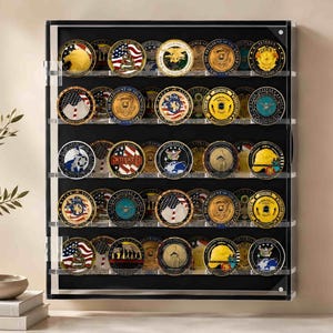 May include: A rectangular black display case with a clear acrylic front door, featuring five horizontal shelves holding a collection of thirty circular military challenge coins. The coins display various colorful emblems, flags, and text.