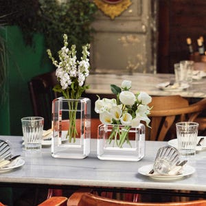 May include: Two clear glass vases with a rectangular design, each holding white flowers. The vases sit on a marble table, alongside glasses and plates. The larger vase contains tall white flowers, while the smaller one holds white calla lilies.