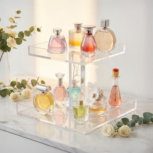May include: A clear, two-tiered acrylic perfume display stand holding various perfume bottles. The bottles are different shapes and sizes, with colorful liquids and decorative caps. Some bottles are round, others square, and some are tall and slender.