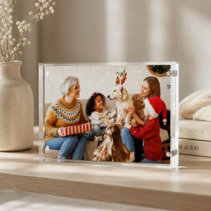 May include: A clear acrylic block photo frame containing a horizontal photograph of a family and a dog wearing reindeer antlers. The frame features magnetic corners and holds a picture of people in holiday sweaters and a red Santa hat.