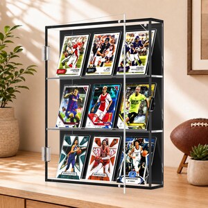 May include: A clear acrylic wall-mounted display case with a black interior frame, featuring three horizontal shelves holding nine sports trading cards. The case has a hinged door with a small latch, showcasing various athlete cards including Carson Palmer, Jay Cutler, Teddy Bridgewater, and others in a grid arrangement.
