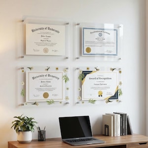 May include: Four framed certificates and awards are mounted on a white wall. Two are plain, while the other two have floral and gold accents. A laptop, plant, and books sit on a wooden desk below.