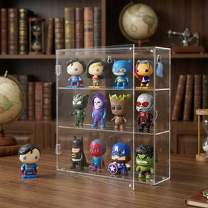 May include: A clear acrylic display case filled with various superhero figurines. The case has multiple shelves and a hinged door. The figurines are brightly colored and depict characters like Superman, Batman, and Groot. The case is on a wooden surface.