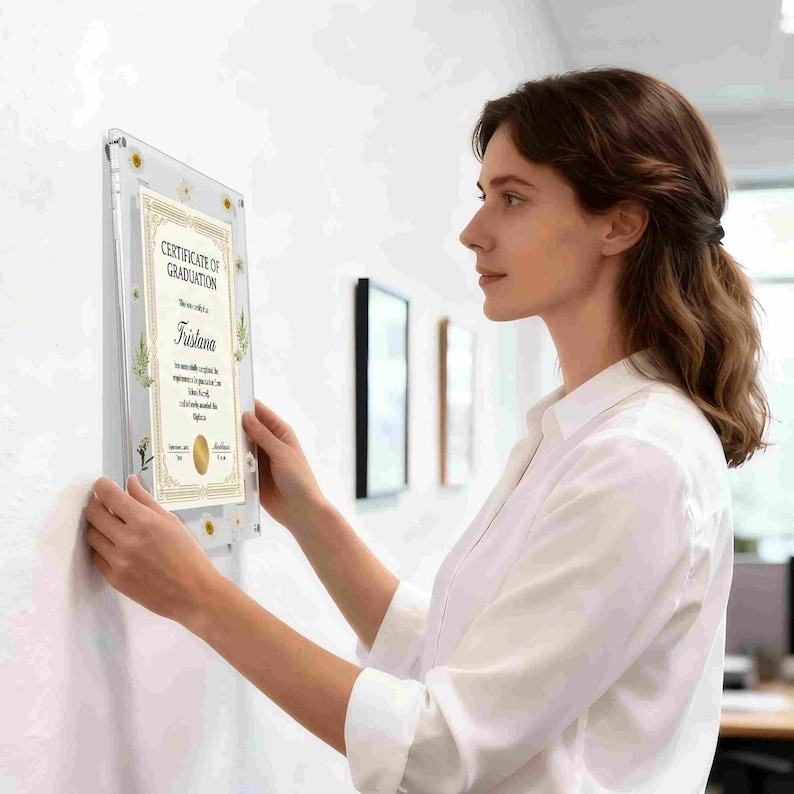 May include: A clear acrylic frame with a graduation certificate. The certificate has gold accents and the words "Certificate of Graduation" at the top. The name "Tristana" is printed on the certificate. The frame is being hung on a white wall.