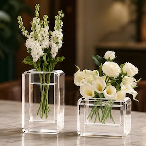 May include: Two clear rectangular acrylic vases with rounded corners. The taller vase holds white snapdragon flowers, while the shorter, wider vase contains a bouquet of white roses and white calla lilies with green stems visible inside.