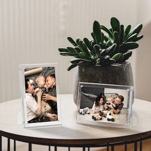 May include: Two clear acrylic picture frames displayed on a table. The left frame is vertical, showing a person holding a toddler. The right frame is horizontal, showing a family playing with toys. A large green succulent plant sits in a gray pot.