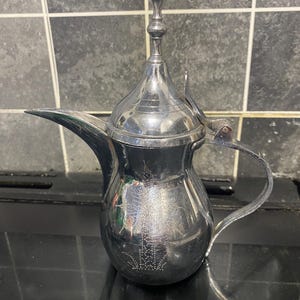 Può includere: Una tazza di ceramica bianca con un manico nero e la scritta "But First, Coffee" in nero, con un carattere a mano. La tazza è su una superficie bianca.