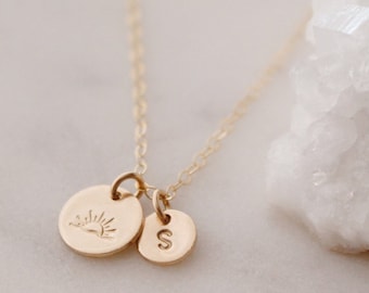May include: A gold necklace with two charms. One charm is a small circle with the letter "S" engraved on it. The other charm is a larger circle with a sun design engraved on it.