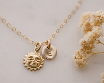 May include: A gold necklace with a sun charm and a small initial charm. The initial charm is a letter E.