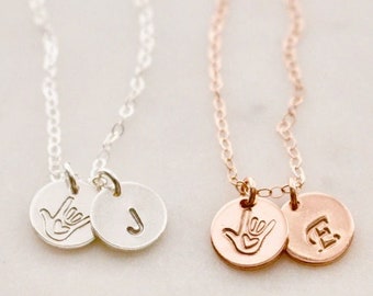May include: Two delicate necklaces with silver and rose gold chains. Each necklace features two charms: a hand sign representing the letter 'I' and a letter charm. The silver necklace has a 'J' charm, and the rose gold necklace has an 'E' charm.
